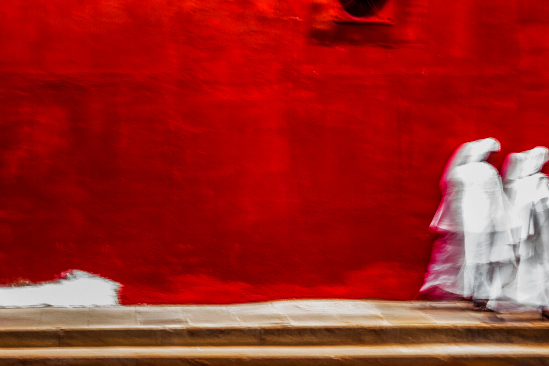 Impressionistic photograph of two women in white cloaks, walking past a red coloured wall in Jeppestown, Johannesburg